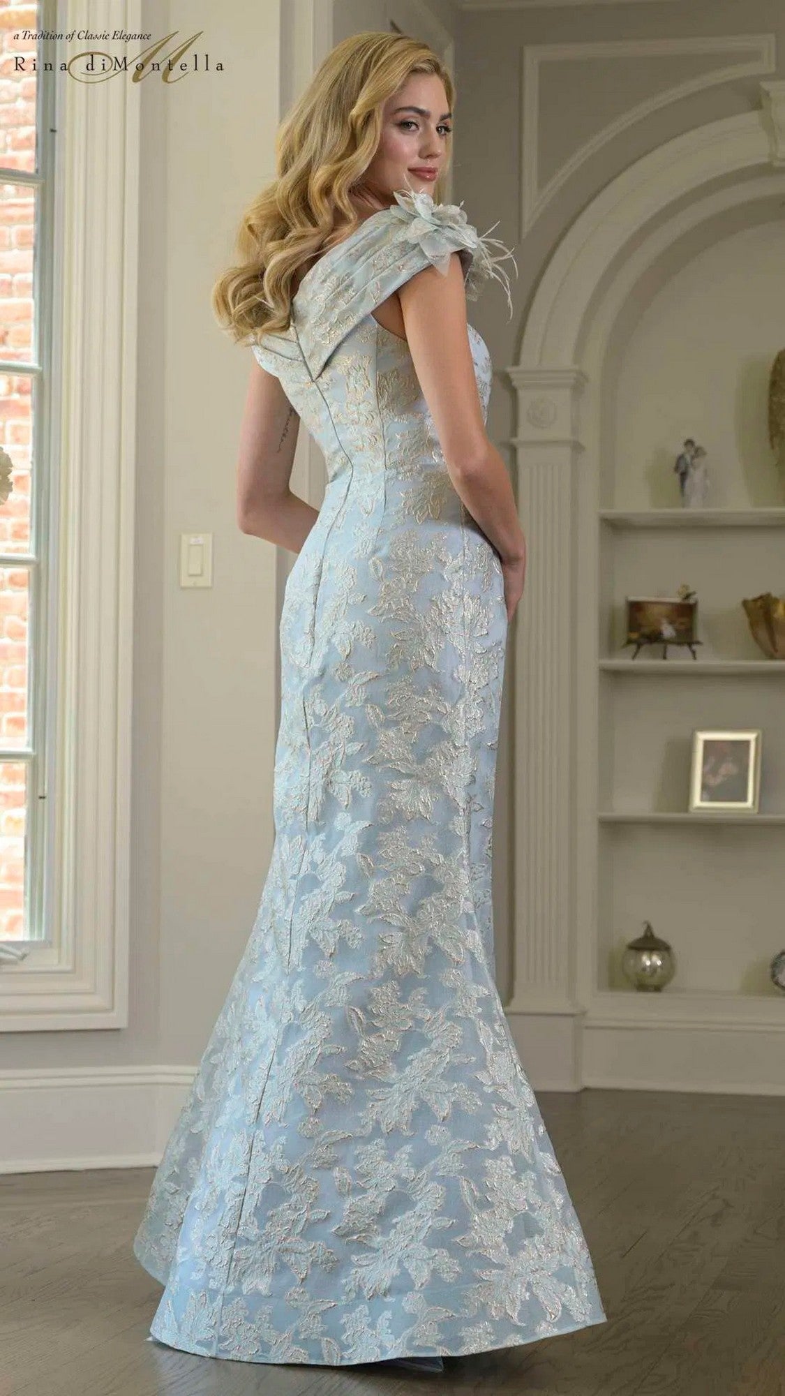 A woman stands indoors facing away from the camera, wearing the Long Formal Dress: Rina Di Montella RD3428 in light blue with a jacquard pattern and off-the-shoulder neckline.